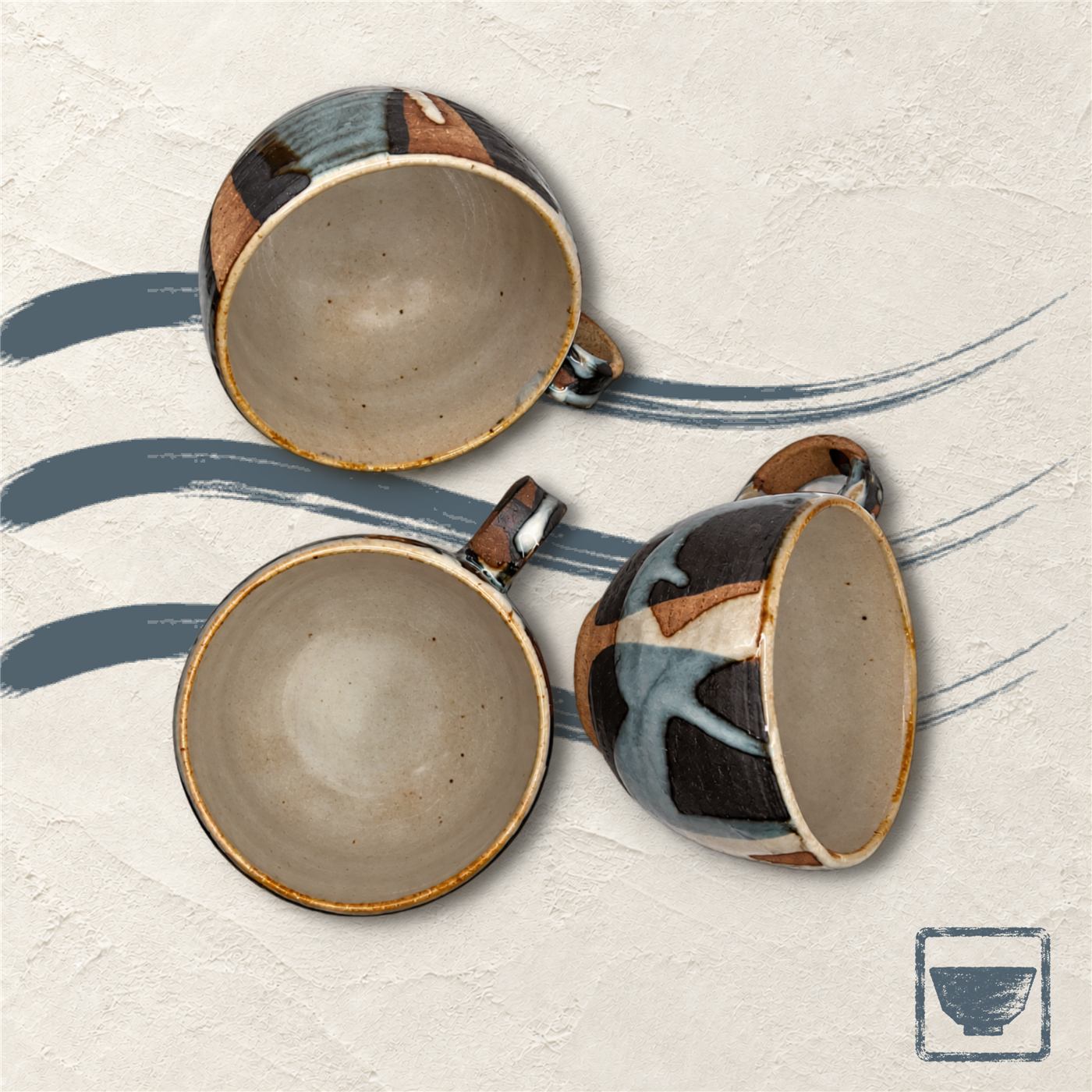 Three stoneware mugs seen from above, their glazes running from earth brown to indigo, on ivory plaster with indigo brush strokes